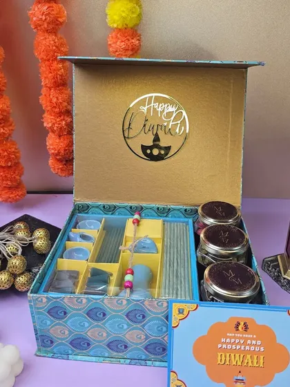 This luxurious Diwali hamper includes a Phool incense kit, jars of dry fruits, and a "Happy Diwali" acrylic cutout, presented in an ornate blue and gold box.