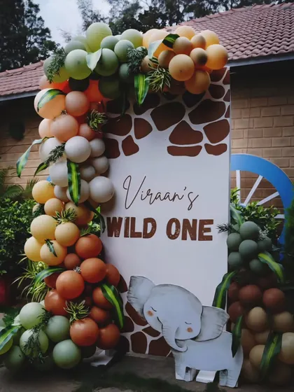 A "Wild One" welcome sign for Viraan's first birthday. The sign is framed by a lush balloon garland in earthy tones of green, orange, and brown, with a cute elephant prop.