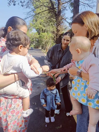 During a nature walk, toddlers and their mothers stop to examine a leaf, showing how learning can happen anywhere.