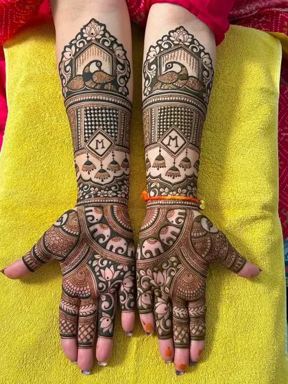 This design features the initial 'M' inside a diamond shape, surrounded by jhumka (earring) and elephant motifs. It's a perfect example of personalized bridal art.