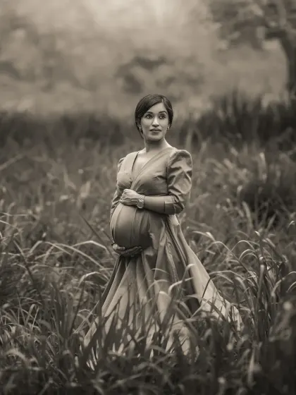 A vintage-inspired black and white outdoor portrait. The expecting mother stands in a field of tall grass, her pose and the soft focus evoking a sense of timeless elegance and quiet strength.