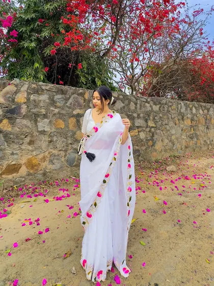 A full-length view of the white mul cotton saree against a stone wall and bougainvillea. The floral embroidery on the saree border complements the natural surroundings perfectly.