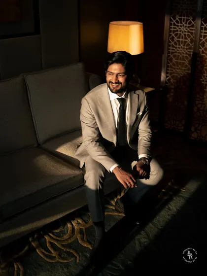 A dapper groom's portrait. The dramatic, directional lighting in the hotel suite creates a moody, cinematic feel, highlighting his sharp suit and confident smile.