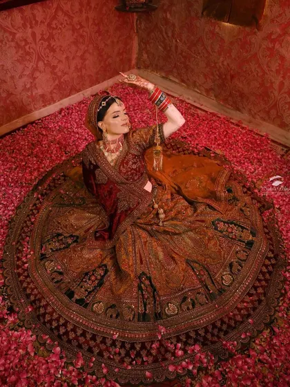 A dramatic, artistic shot of a bride in a deep maroon lehenga, surrounded by rose petals. This shows the cinematic quality our outfits can bring to your wedding photos.