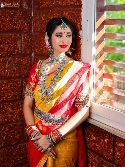The bride stands by a window, bathed in natural light. The sun highlights the beautiful colors of her yellow and red silk saree and the intricate details of her silver jewelry.