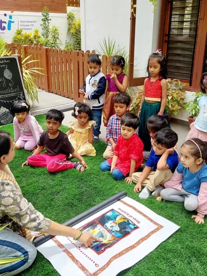 Children sit on the grass, captivated by a story about Janmashtami. Celebrating festivals outdoors connects our cultural learning with the natural environment.