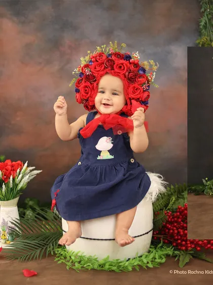 A happy baby girl wearing a stunning red rose bonnet, sitting in a setup with lush greenery.