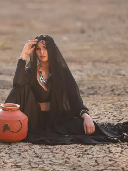 A conceptual photoshoot featuring a model in a simple yet elegant black lehenga. The outfit is styled with a sheer dupatta and traditional jewelry for a rustic, powerful look.