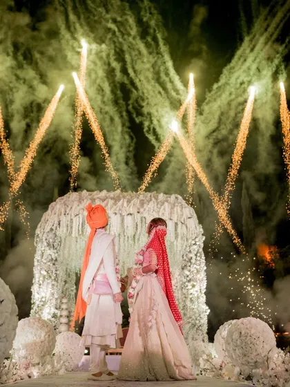 A moment frozen in time, where love and light collide. The couple stands before a floral mandap as fireworks paint the sky, a signature of my grand wedding planning.