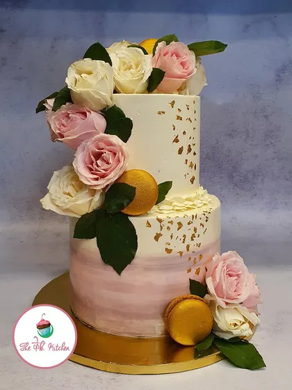 This two-tiered cake combines a soft pink watercolor effect with a rustic buttercream finish. It's decorated with fresh roses and gold macarons for an elegant touch.