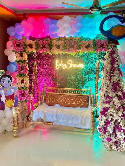 An indoor baby shower setup with a Radha-Krishna theme, featuring a decorated swing, character cutouts, and a colorful balloon arch.