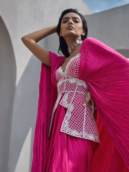 A vivid, hot pink ensemble featuring an embellished corset top paired with a draped skirt and a dramatic, crushed fabric cape. The look is a play on structure and fluidity.