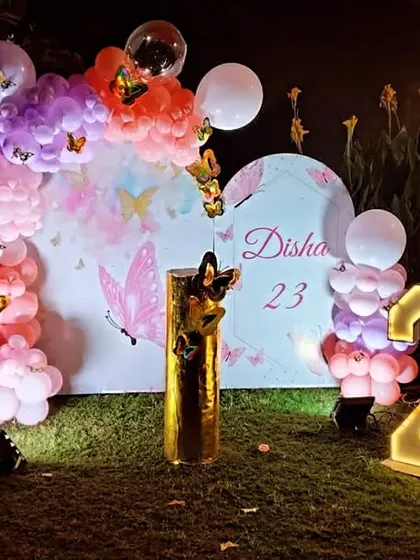 An outdoor butterfly-themed setup for a 23rd birthday, featuring a balloon arch, marquee numbers, and a custom backdrop.