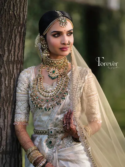 A close-up portrait of a bride in her white and gold saree. The makeup is kept soft and luminous to let the stunning emerald-studded temple jewellery stand out.