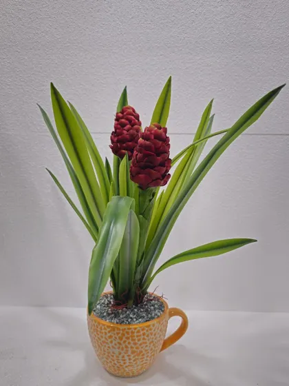 A unique artificial plant with red cone-like flowers and long green leaves, presented in a fun, mug-shaped yellow pot.