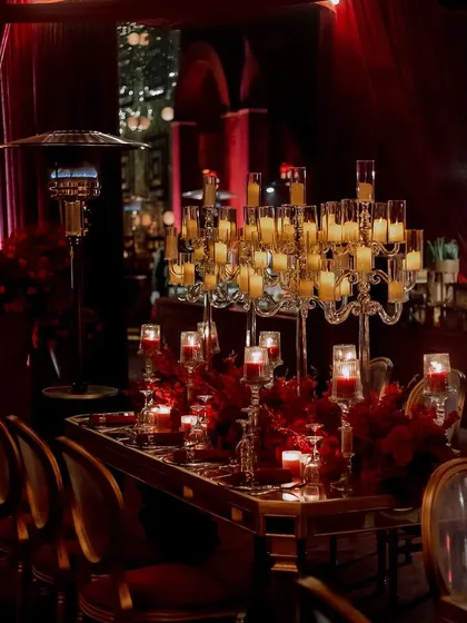 A long shot of the dining tables, featuring towering candelabras and lush red floral arrangements. The mirrored tabletops reflect the candlelight, amplifying the warm and opulent feel of the room.