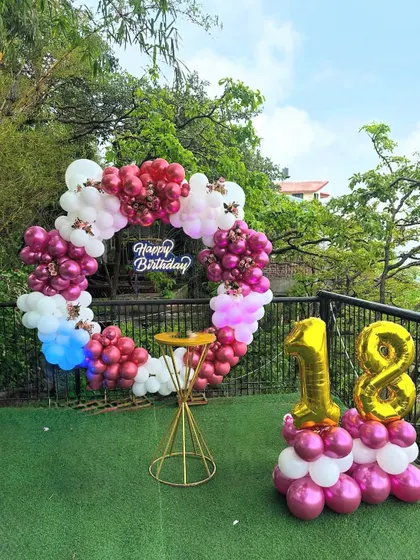 An outdoor 18th birthday party decoration on a terrace. The setup includes a circular balloon wreath in pink and white, a neon sign, and a balloon pillar with the number '18'.