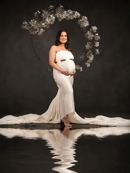 An elegant portrait against a dark background, framed by a silver floral hoop. The simple white gown and water reflection create a sophisticated and artistic composition.