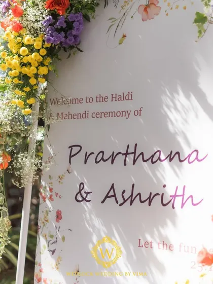 A vibrant floral backdrop and welcome sign for a colorful Haldi and Mehendi ceremony. The mix of orange, blue, purple, and yellow flowers created a stunning photo opportunity for the couple and their guests.