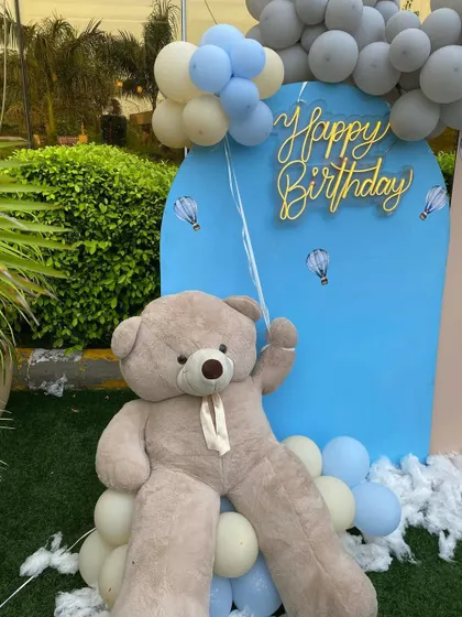 A giant, cuddly teddy bear holds a balloon bunch next to the neon "Happy Birthday" sign, adding a playful and adorable touch to the decor.