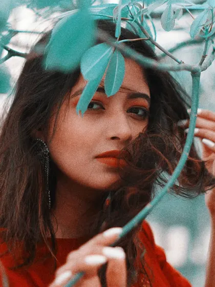 A close-up portrait with a creative twist, using leaves to frame the model's face. The teal and red color grading gives the image a unique, artistic feel while drawing attention to her direct gaze and the simple beauty of the natural elements.