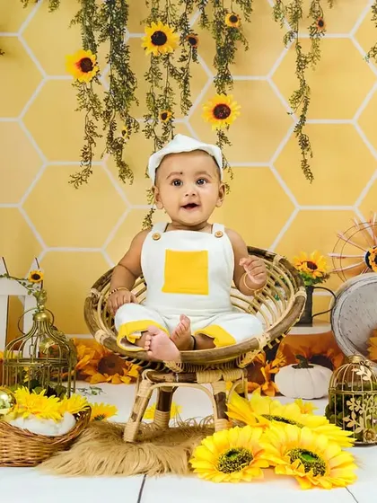 A baby boy in a white and yellow outfit sits happily in a sunflower-filled scene.
