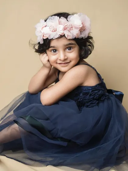 A little girl in a navy blue dress and a floral crown, looking at the camera with a sweet, knowing smile.
