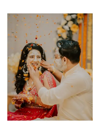A sweet moment where the groom gently applies haldi to the bride's face, with flower petals falling around them.