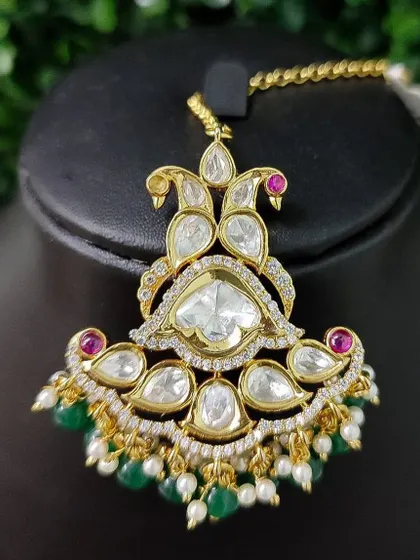 An exquisite kundan maang tikka with a unique paisley design. The piece is accented with small rubies and green bead drops for a regal look.