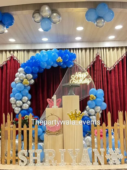 A blue and silver balloon ring with a 'Happy Birthday' sign, set on a stage with a red curtain backdrop.