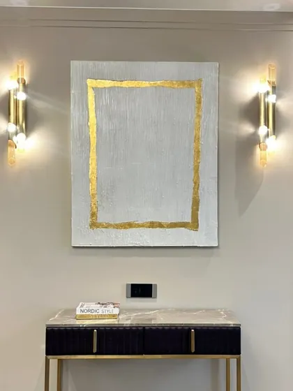 A beautiful art installation in a Hyderabad home. This piece features a simple, elegant design with a textured white background and a border of 24ct gold leaf, perfectly framed by the wall sconces.