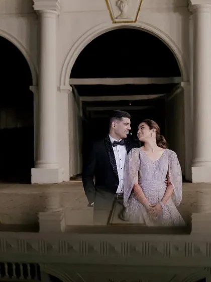 A wide shot of a couple during their pre-wedding or reception shoot, set against a grand, arched building at night. The composition creates a sense of drama and romance.