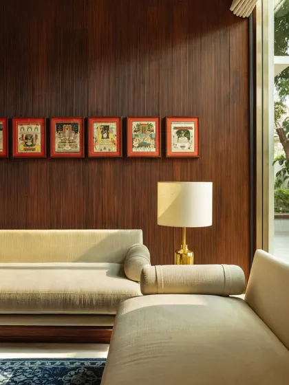 Sunlight grazes a wood-paneled wall adorned with a series of traditional paintings, illuminating a modern sofa. This composition highlights our approach of integrating art and heritage into contemporary living spaces.