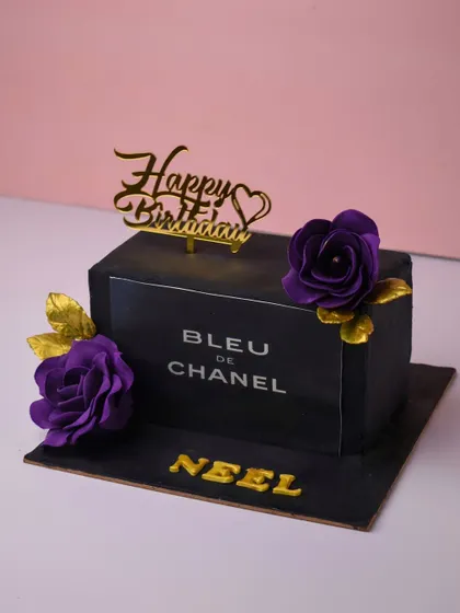 A chic cake designed to look like a Bleu de Chanel perfume bottle. This rectangular cake is covered in black fondant with silver lettering and adorned with deep purple sugar flowers and gold leaves for a touch of luxury.