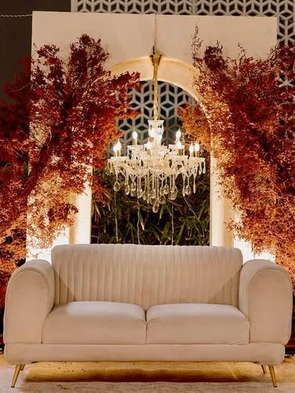 The reception stage, where a simple white sofa is set against a backdrop of arched panels, crystal chandeliers, and dramatic bursts of red foliage, creating a look of modern romance.