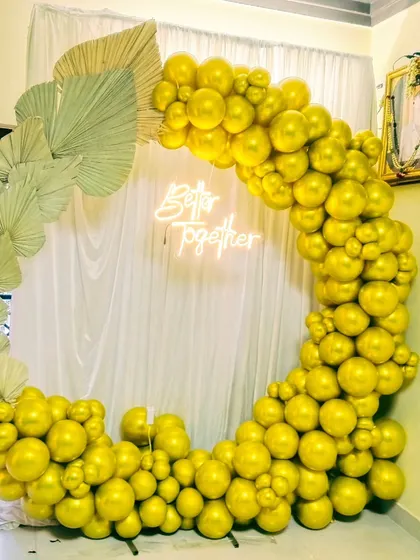 An elegant engagement or anniversary setup with a 'Better Together' neon sign. The ring is decorated with solid gold-colored balloons and dried palm leaves.