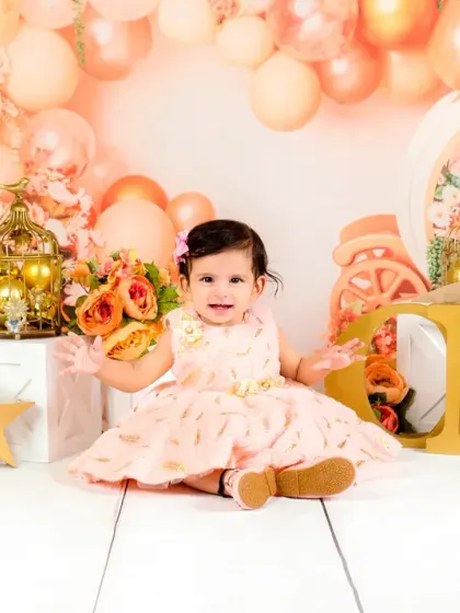 A baby girl in a beautiful peach dress sits in front of "ONE" blocks during her first birthday photoshoot, surrounded by matching balloons.