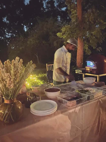 An evening pizza party in full swing. My chef works at the prep table under ambient lighting, with the glow of the wood-fired oven behind him, creating a cozy and inviting atmosphere for guests.