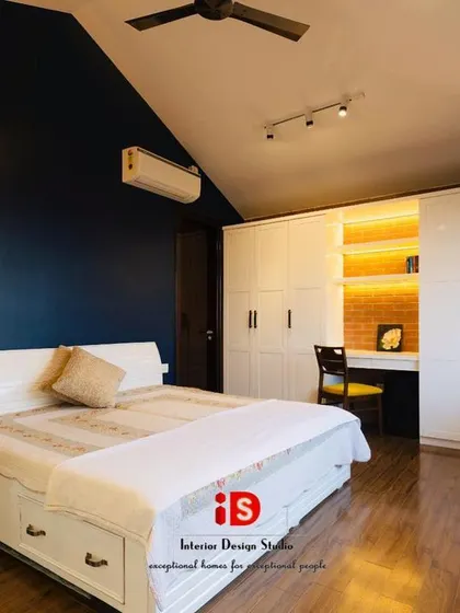 This bedroom design combines rustic and modern elements for a unique feel. A deep blue accent wall contrasts with an exposed brick wall, while the white bed with built-in storage and a sleek wardrobe keep the look clean and functional.