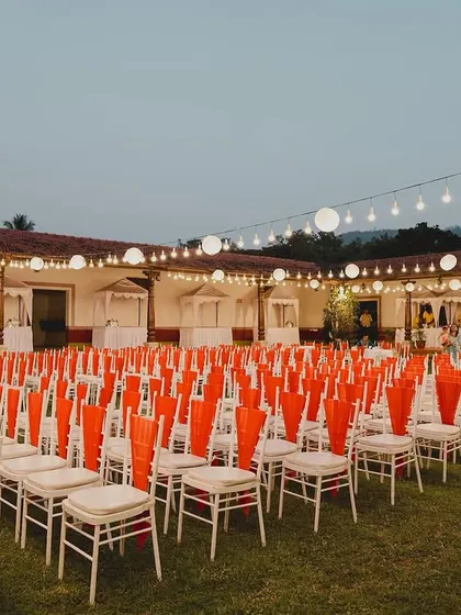 The Sangeet seating arrangement as night falls. The combination of white chairs with bright orange accents and overhead lanterns creates a festive and inviting atmosphere for guests.