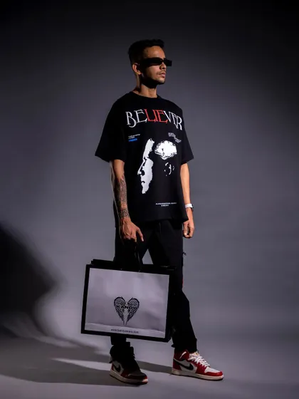 A full-length studio shot of a male model in an oversized graphic tee and sneakers, holding a branded bag. This is a complete look that's perfect for a lookbook, showing how different brand items can be styled together.