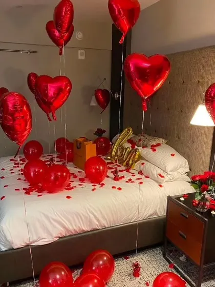 A classic romantic room setup with red heart-shaped balloons, rose petals scattered on the bed and floor, and 'Love' foil balloons. A bouquet of fresh roses on the side table completes the look.