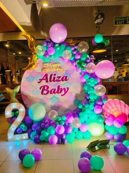 A mermaid-themed second birthday party with a backdrop shaped like a mermaid scale pattern. The balloon colors are purple, teal, and green, with a large marquee number '2'.