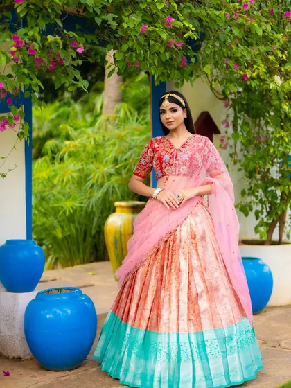 A stunning half saree with a unique color combination of pink, orange, and turquoise. The vibrant blue pots in the background create a beautiful, artistic composition.