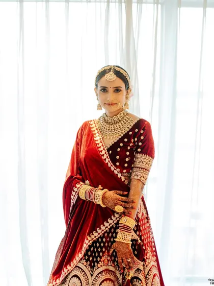 A classic bridal portrait against a bright window, her red velvet lehenga and intricate jewelry creating a stunning and timeless look.
