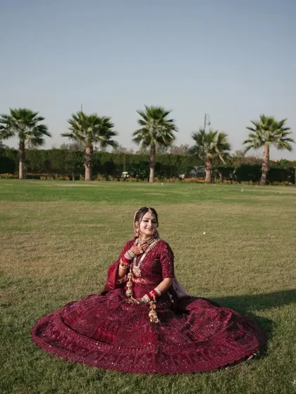 A stunning outdoor bridal portrait. The bride sits gracefully on the lush green lawn, her maroon lehenga spread beautifully around her.