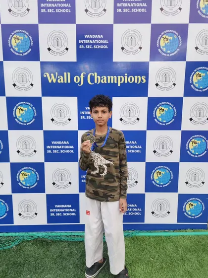A student standing proudly in front of the "Wall of Champions" at the CBSE Taekwondo Championship 2023.