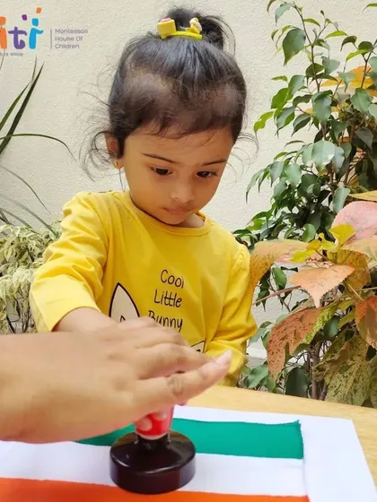 A young child carefully uses a stamp to place the Ashoka Chakra on her flag. This action develops her fine motor control and understanding of our national symbol.
