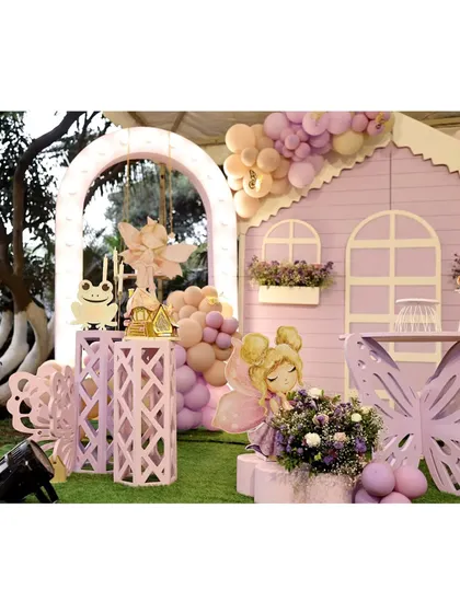 An enchanting outdoor fairy party setup. A life-sized purple fairy house is the main feature, complemented by a light-up arch, whimsical frog and fairy props, and beautiful floral arrangements.