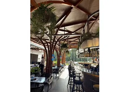 The tree-like support columns and hanging greenery create a forest-like canopy effect within the bar area. This is a prime example of biophilic design that mimics natural patterns to create a calming and restorative environment.